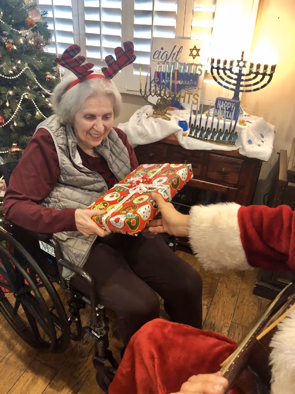 An elderly woman in a wheelchair wearing reindeer antlers receives a wrapped Christmas gift from a person dressed in a Santa Claus outfit. Behind them is a decorated Christmas tree and a table with Hanukkah menorahs and a sign that says 'eight nights Happy Hanukkah'.