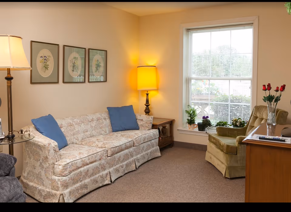 Cozy living room with a floral sofa, armchair, lamps, framed botanical prints, and a window ledge with potted plants.