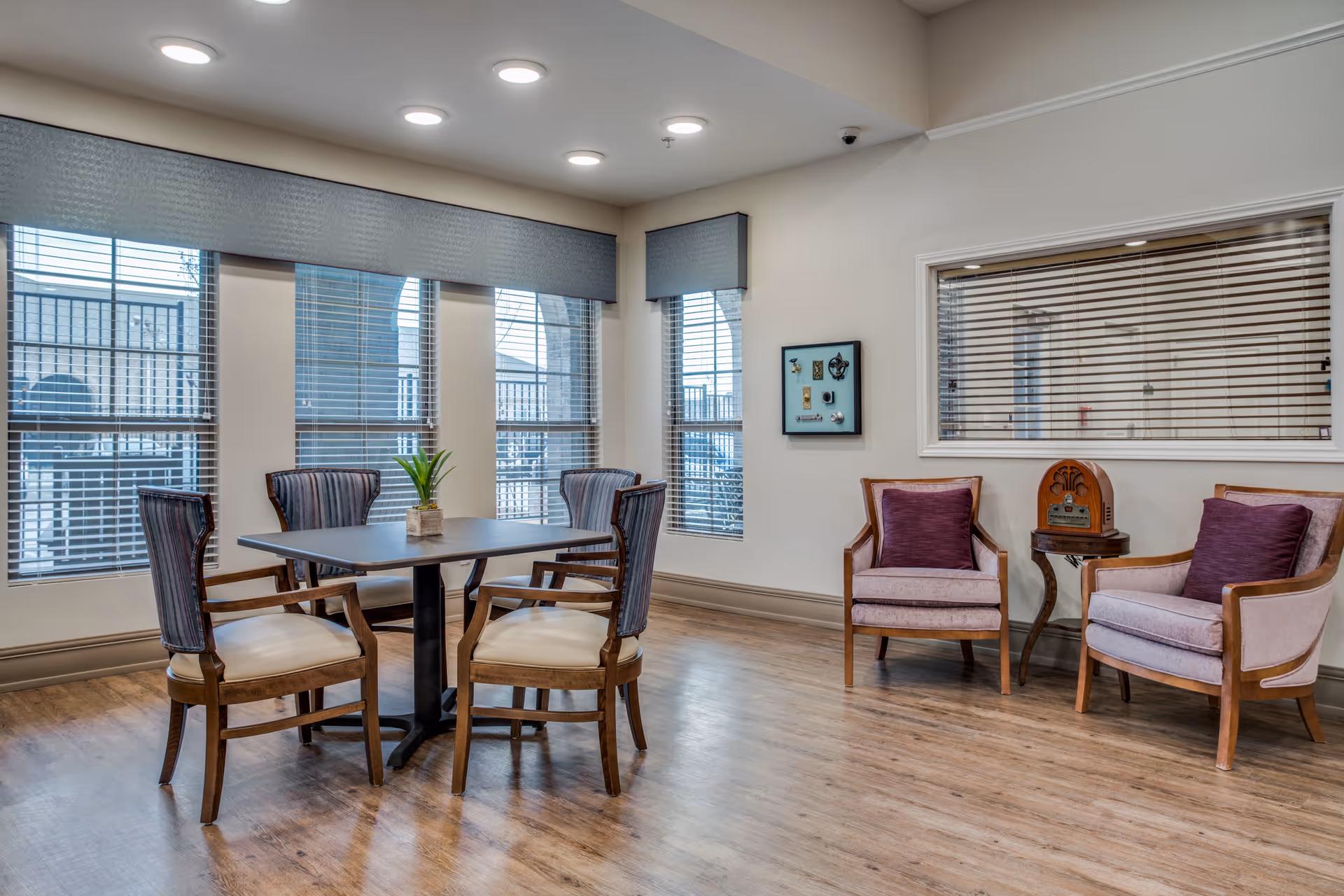 A well-lit sitting area in a senior living facility with a square table surrounded by four wooden chairs with cushioned seats and striped backs. To the right, there are two wooden armchairs with purple cushions and a small round table between them holding a vintage-style radio. Large windows with blinds and valances let in natural light, and the floor is wood.