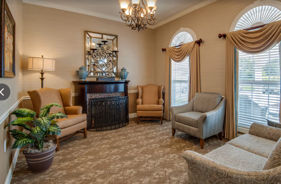Sunlit sitting room with upholstered armchairs, a small fireplace, chandelier, and arched windows with drapes.