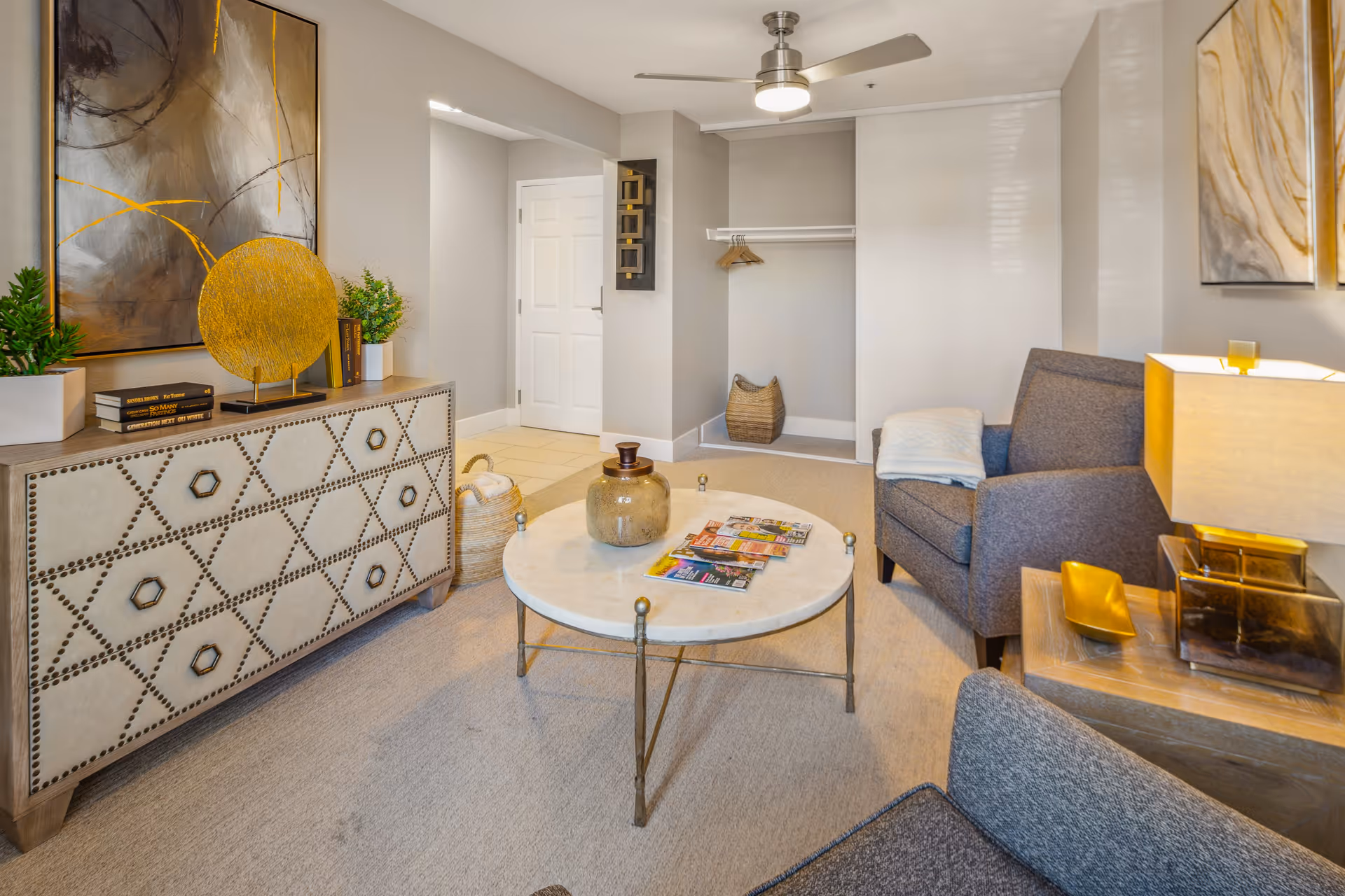 A cozy living room area with two gray armchairs, a round marble coffee table with magazines, a decorative sideboard with plants and art pieces, a lamp on a wooden side table, and a ceiling fan. The walls are painted light gray and there is an open closet space with a basket and hangers.
