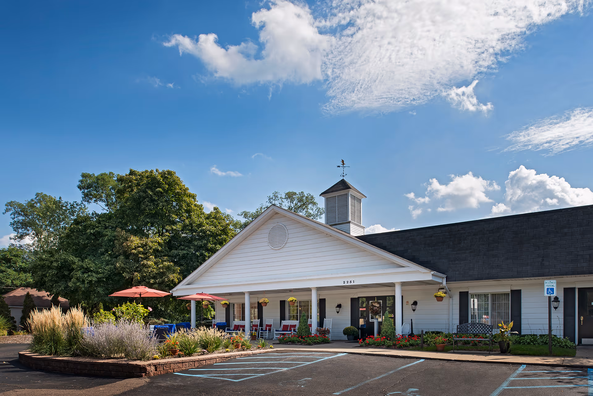 Exterior view of a single-story white building with a black roof, featuring a covered porch with red chairs and tables with red umbrellas. There are plants and flowers in front of the building, a parking lot with handicap parking spaces, and trees in the background under a partly cloudy blue sky.