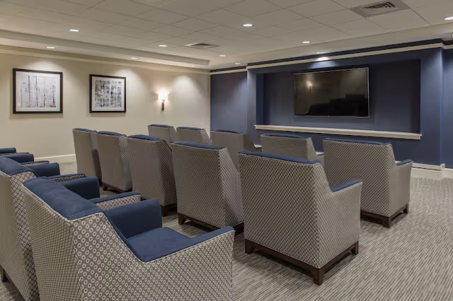 A small theater-style room with two rows of patterned armchairs facing a large flat-screen TV mounted on a dark blue accent wall. The room has beige walls with two framed black and white artworks and wall sconces providing soft lighting. The ceiling has recessed lights and a white grid pattern.