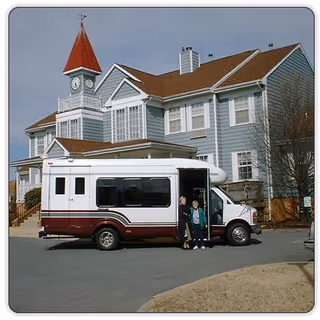 A maroon-and-white shuttle bus parked in front of a large multi-story retirement building with two people standing at the bus doorway.