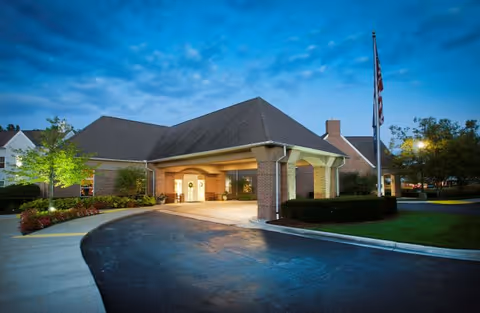 Exterior view of a senior living facility building at dusk with a covered entrance, brick walls, a flagpole with an American flag, and surrounding landscaping including trees and bushes.