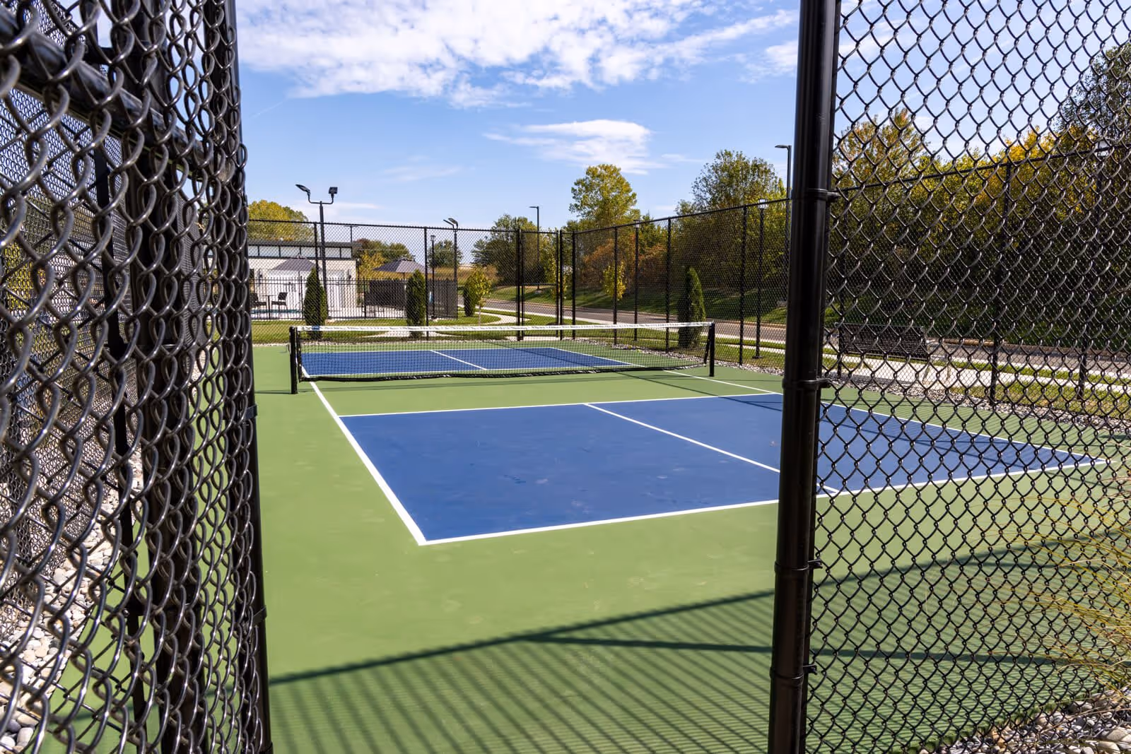 Outdoor fenced pickleball court with a blue and green playing surface under a partly cloudy sky, surrounded by trees and a pathway.
