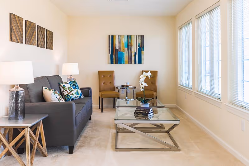 A bright living room with a gray sofa adorned with patterned pillows, two brown chairs, a glass coffee table with books and a white orchid, two side tables with lamps, and abstract wall art. Large windows with blinds allow natural light to fill the space.