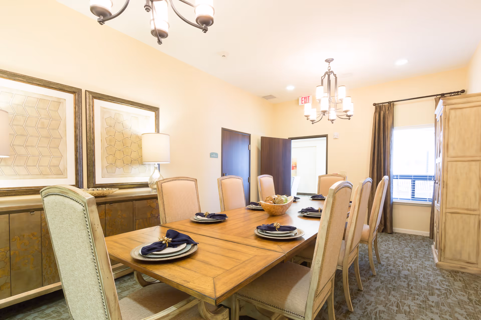 A well-lit dining room with a long wooden table set for eight people. Each place setting includes a plate, a dark blue napkin with a gold napkin ring, and silverware. The room features beige walls, two framed geometric art pieces, a sideboard with a lamp, a window with dark curtains, and two chandeliers hanging from the ceiling.