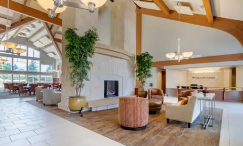 Spacious and well-lit senior living facility common area with high vaulted ceilings and wooden beams. The room features a large stone fireplace in the center, surrounded by comfortable seating including armchairs and a small round table. There are large potted plants near the fireplace, and in the background, a dining area with tables and chairs is visible. The reception desk with the sign 'Woodlands Creek' is also seen on the right side of the image.