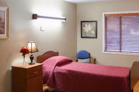 A bedroom in a senior living facility with burgundy bedspread, nightstand, lamp, and window.