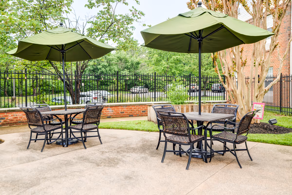 Outdoor patio area with two round tables, each surrounded by four wicker chairs and shaded by large green umbrellas. The patio is paved and bordered by a brick wall and black metal fence, with trees and greenery in the background.