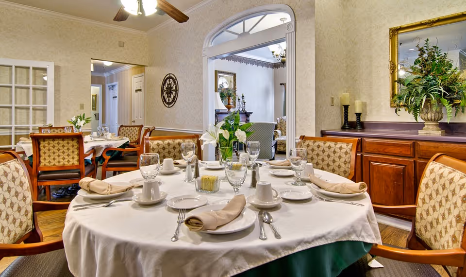 A formal dining room set for a meal with a round table, place settings, upholstered chairs, and a sideboard with plants and decor.
