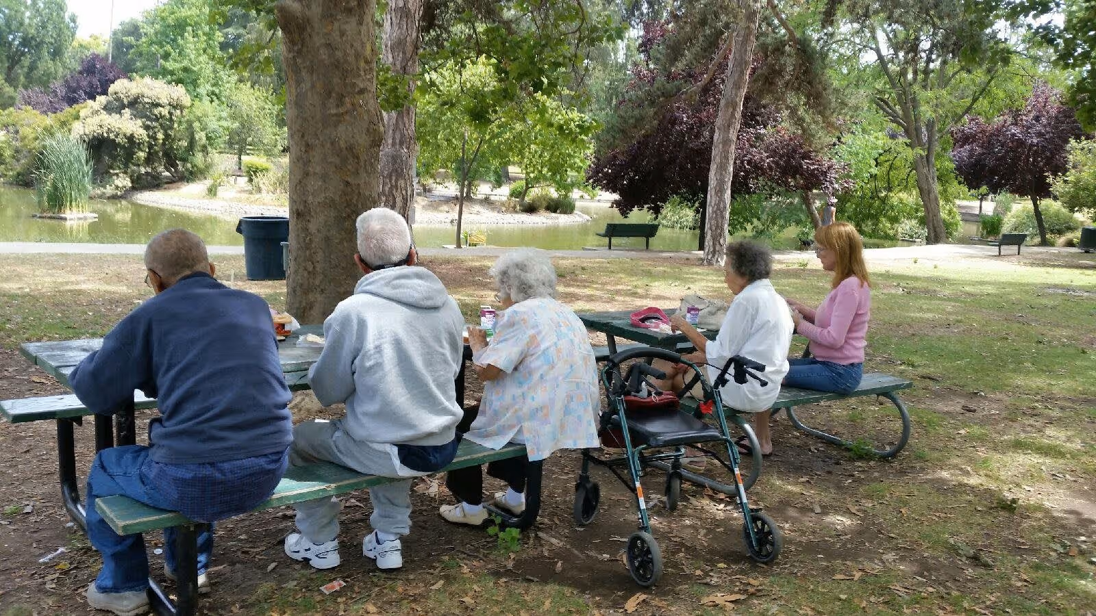 Five people sitting at a green picnic table in a park near a pond. Four elderly individuals and one younger woman are seated on benches, with a walker placed next to one of the elderly women. Trees and greenery surround the area, creating a peaceful outdoor setting.