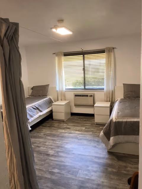 A simple, clean bedroom with two single beds placed on opposite sides of the room. Each bed has gray bedding and a pillow. Between the beds are two white nightstands, and a window with blinds and sheer curtains is centered on the far wall. Below the window is a wall-mounted air conditioning unit. The floor has a wood-like finish, and the walls are painted white.