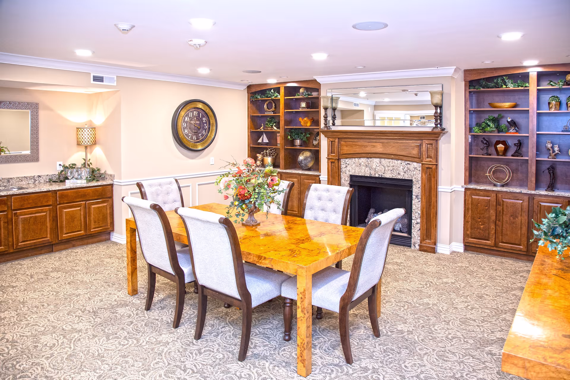 A cozy dining room with a wooden table surrounded by six cushioned chairs. The room features a decorative fireplace with a large mirror above it, built-in wooden shelves with various decorative items, a wall clock, and a sideboard with a lamp and plants. The carpet has a patterned design and the walls are painted beige.