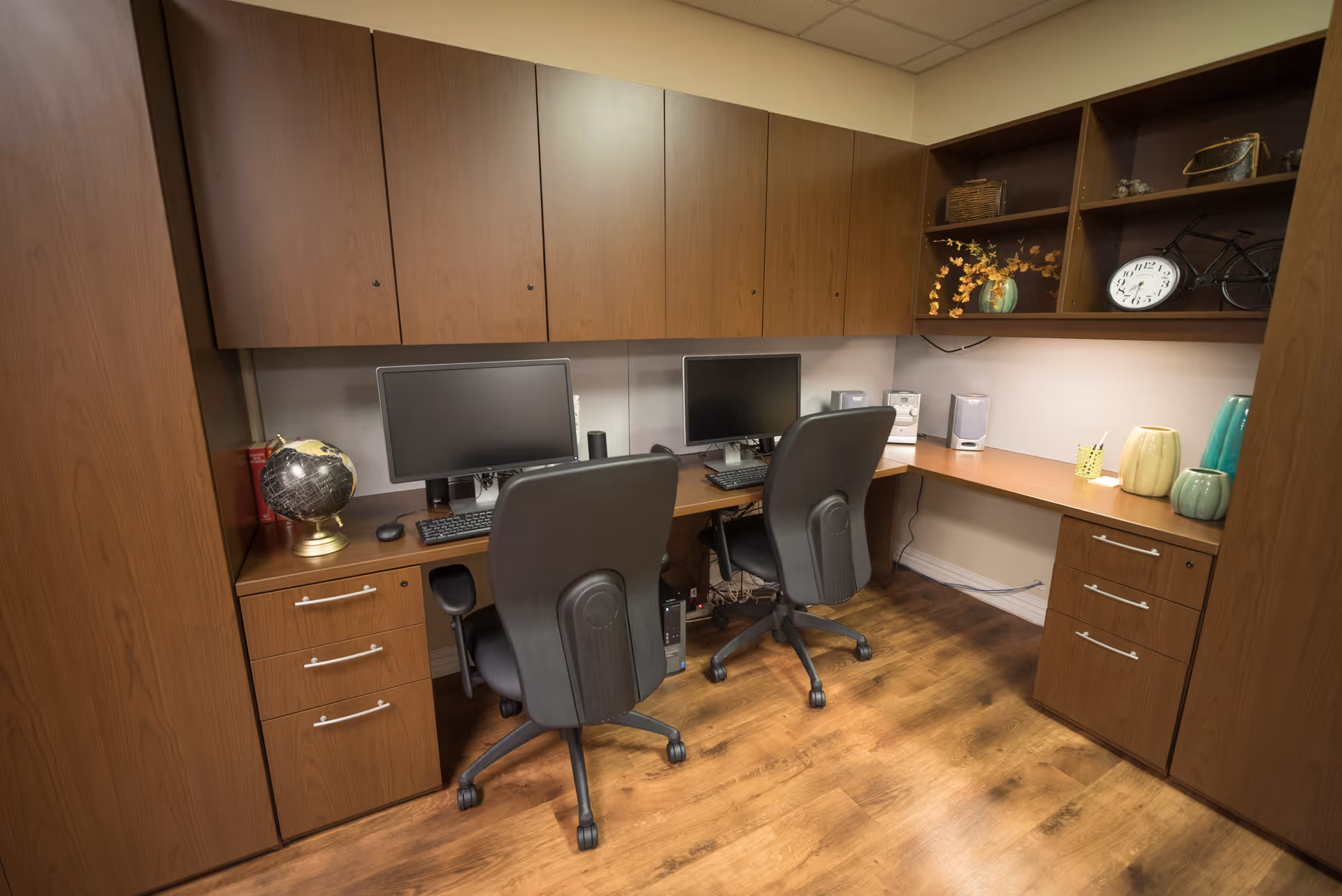 Two computer workstations with office chairs under wood cabinets and shelving in a small interior office area.