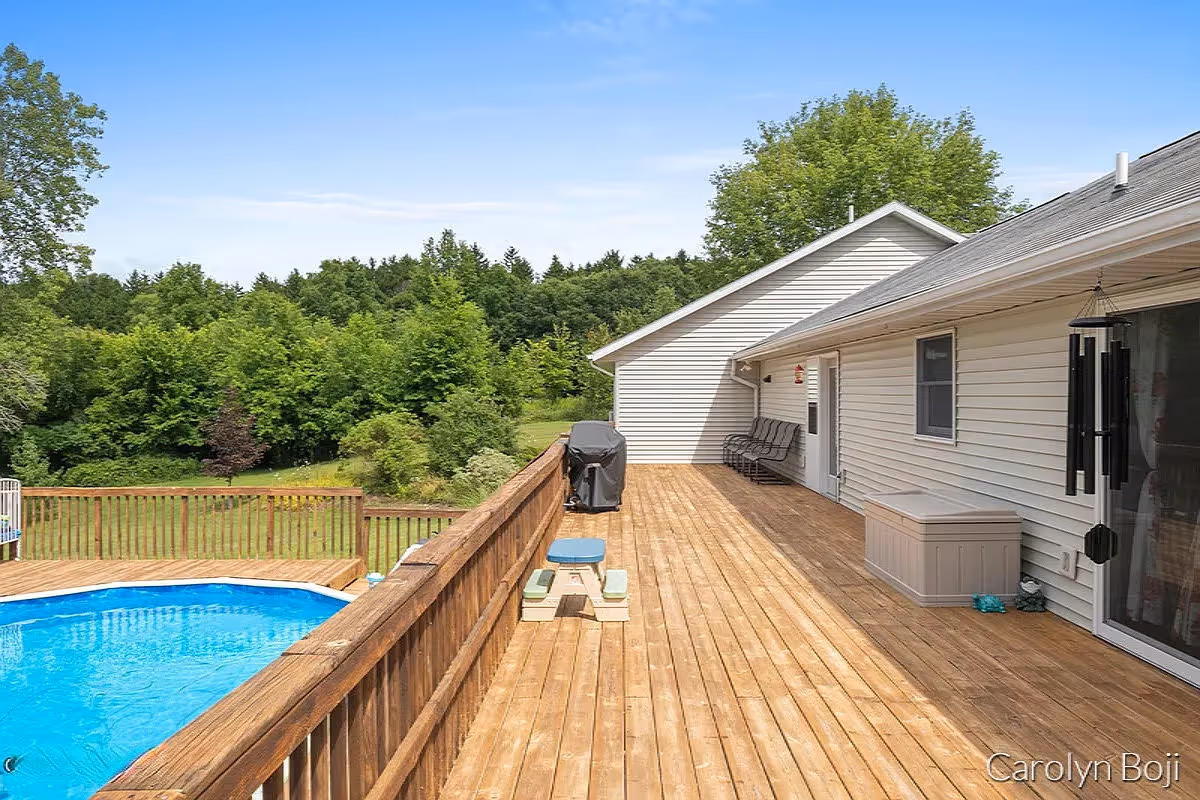 A spacious wooden deck attached to a light-colored house with a railing overlooking a blue above-ground swimming pool. The deck has a small picnic table, a covered grill, a storage box, and a bench near the house wall. In the background, there are green trees and a clear blue sky.