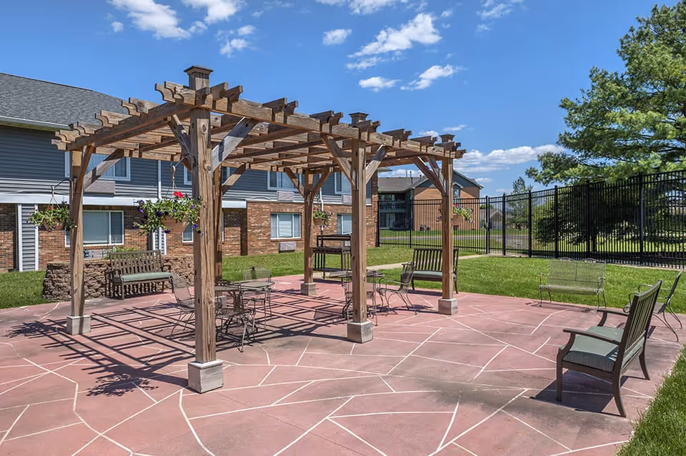 Outdoor patio area with a wooden pergola structure, metal chairs and tables, benches with cushions, hanging flower pots, and a barbecue grill. The patio is surrounded by grass, a black metal fence, and residential buildings under a blue sky with scattered clouds.