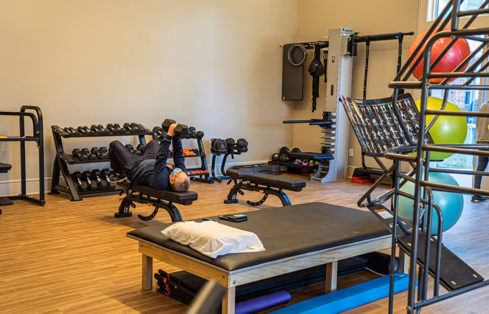 An elderly man wearing a mask is lying on a workout bench lifting dumbbells in a fitness room equipped with various weights, exercise balls, and workout equipment. The room has wooden flooring and light-colored walls with a large window letting in natural light.
