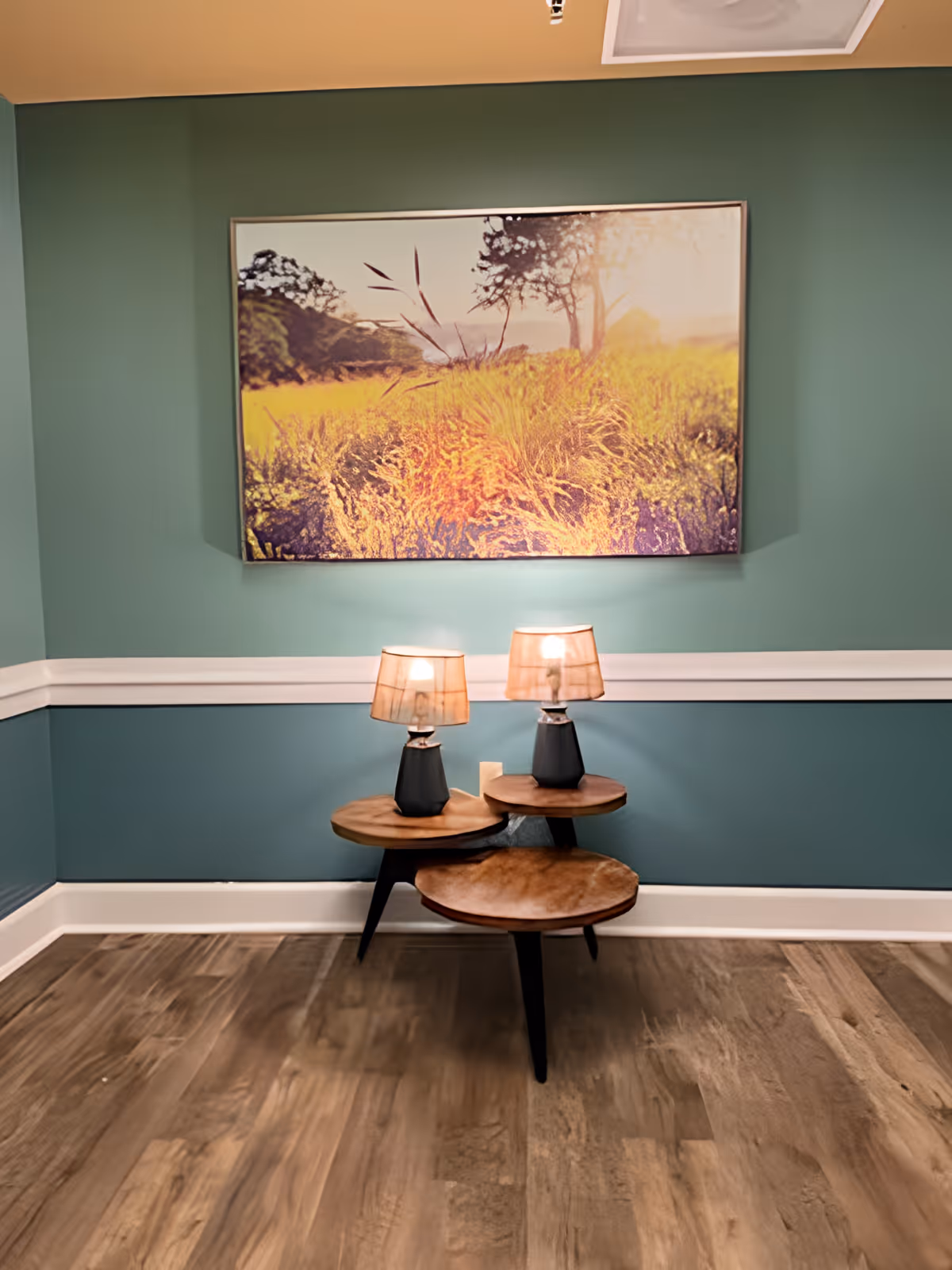A corner of a room with teal walls and white trim, featuring a wooden floor. There is a three-tiered wooden side table with two small lamps on the top tiers. Above the table hangs a large framed picture of a sunlit field with trees in the background.