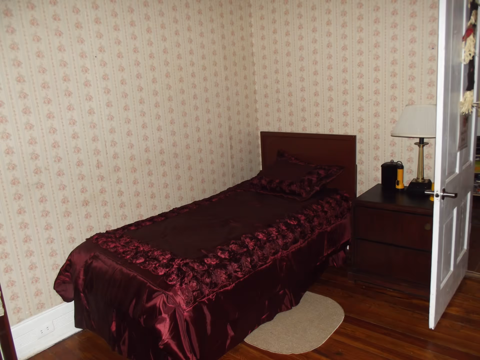A small bedroom with a single bed covered in a dark red satin bedspread with decorative ruffles. The bed is positioned against a wall with floral patterned wallpaper. Next to the bed is a dark wooden nightstand with a lamp, a flashlight, and some other small items. The floor is wooden with a small oval rug beside the bed. The door to the room is open.