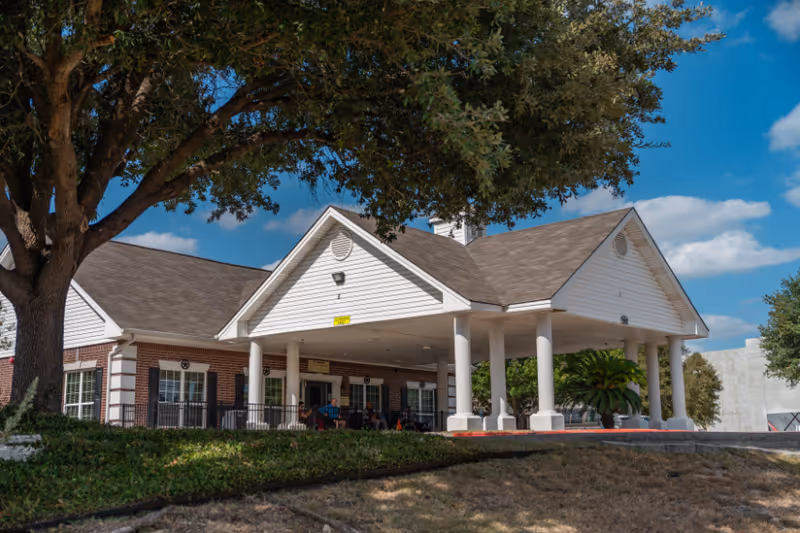Front entrance of a nursing and rehabilitation building with a covered portico supported by columns, a porch with people seated, and a large tree in front.