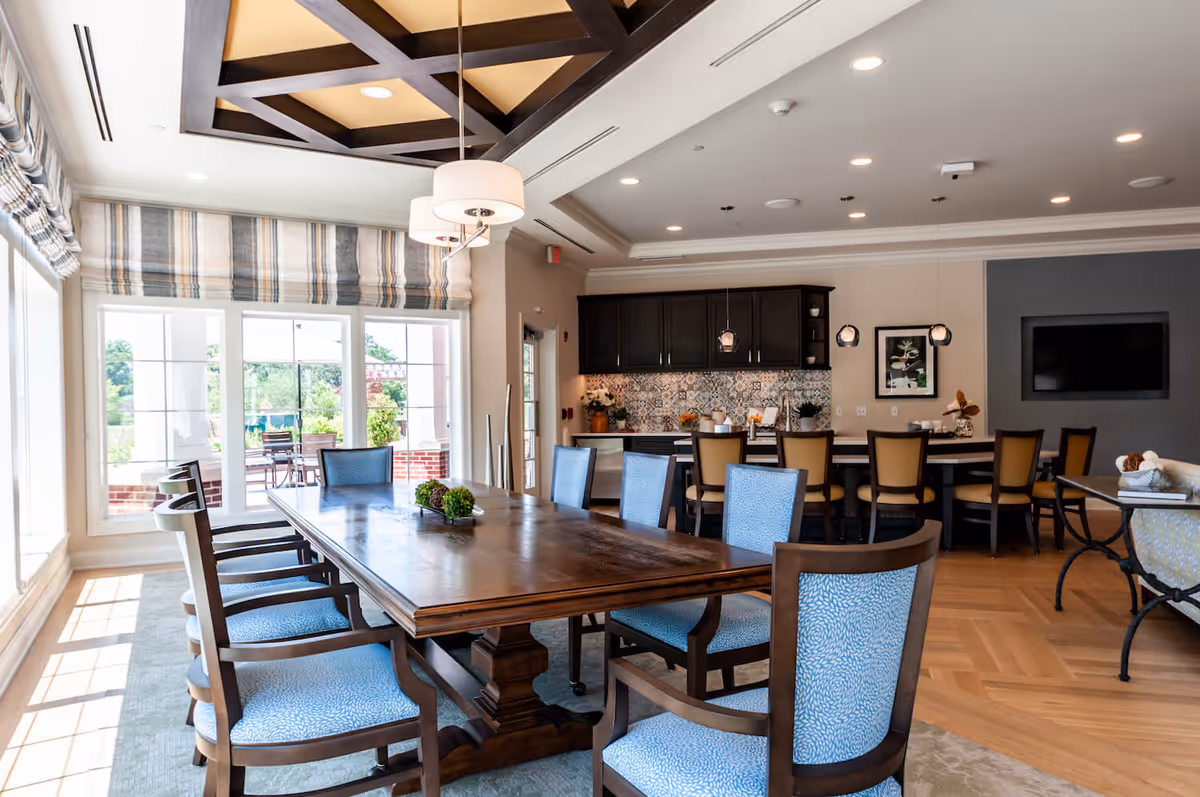 A bright and spacious dining area in Arbor Terrace Mount Laurel featuring a large wooden dining table surrounded by blue upholstered chairs. The room has large windows with striped curtains allowing natural light to fill the space. In the background, there is a kitchen area with dark cabinets, a patterned backsplash, and a long counter with several chairs. The ceiling has decorative wooden beams and modern pendant lights hang above the kitchen counter.