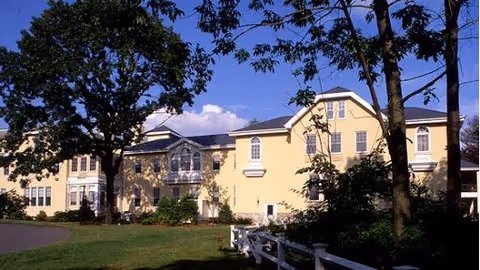 Exterior view of a large, yellow senior living facility building with multiple windows, surrounded by trees and greenery under a clear blue sky.
