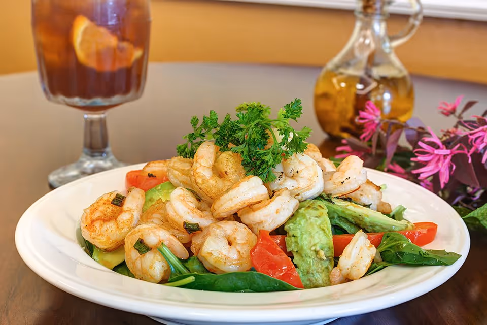 A plate of shrimp salad with spinach, avocado slices, and tomato wedges, garnished with a sprig of parsley. In the background, there is a glass of iced tea with lemon and a glass bottle with a stopper, along with some pink flowers on a wooden table.