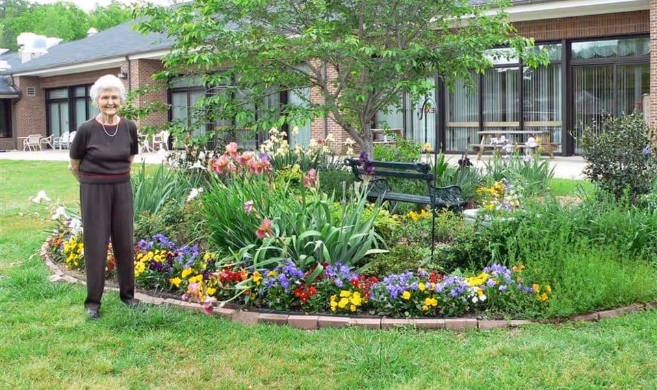 An elderly woman standing on grass next to a colorful flower garden with various blooming flowers and a small tree. Behind the garden is a brick building with large windows and outdoor seating.