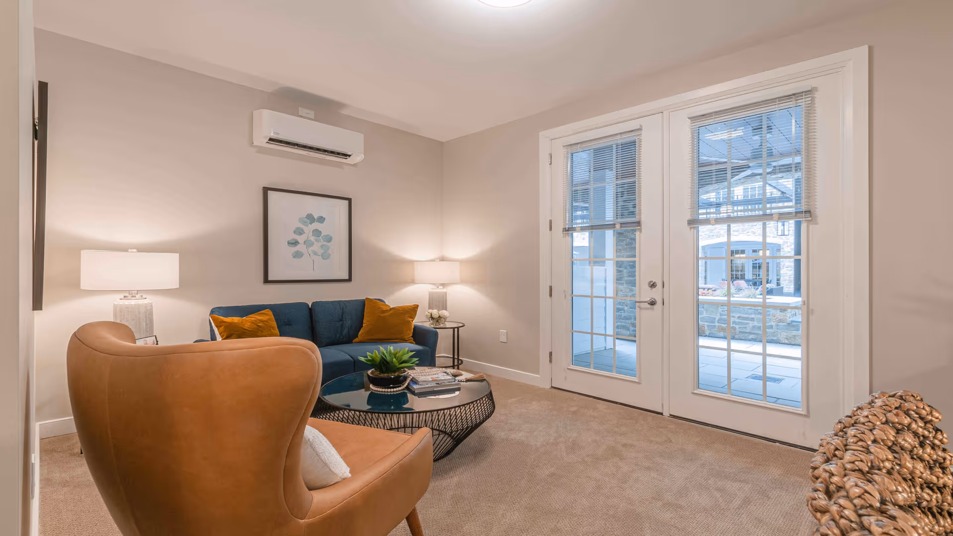 A cozy living room with a blue sofa adorned with orange pillows, a brown leather armchair, a round glass coffee table with a plant and books, two table lamps on either side of the sofa, and double glass doors leading to an outdoor area.