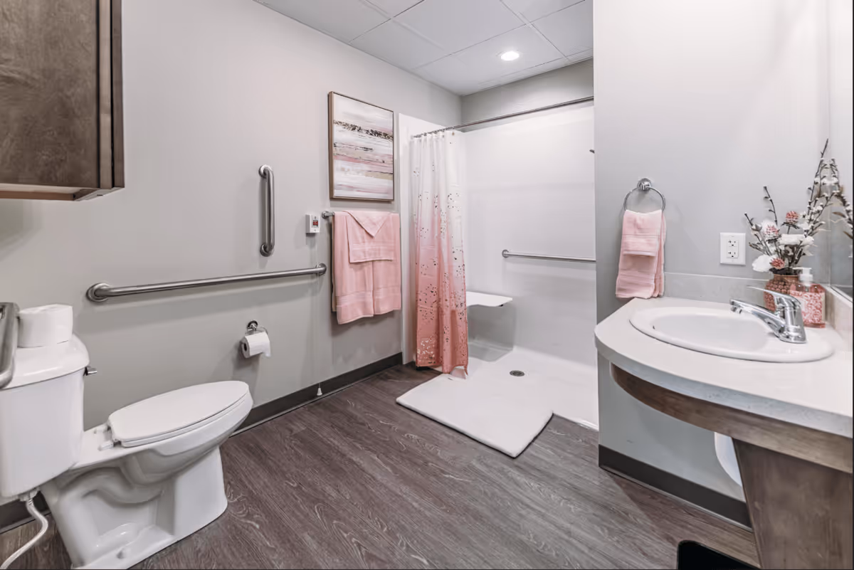 A clean and accessible bathroom with a white toilet, grab bars on the walls, a shower area with a pink and white shower curtain, a white shower seat, a white bath mat, a sink with a countertop, pink hand towels, and a decorative vase with flowers on the counter.