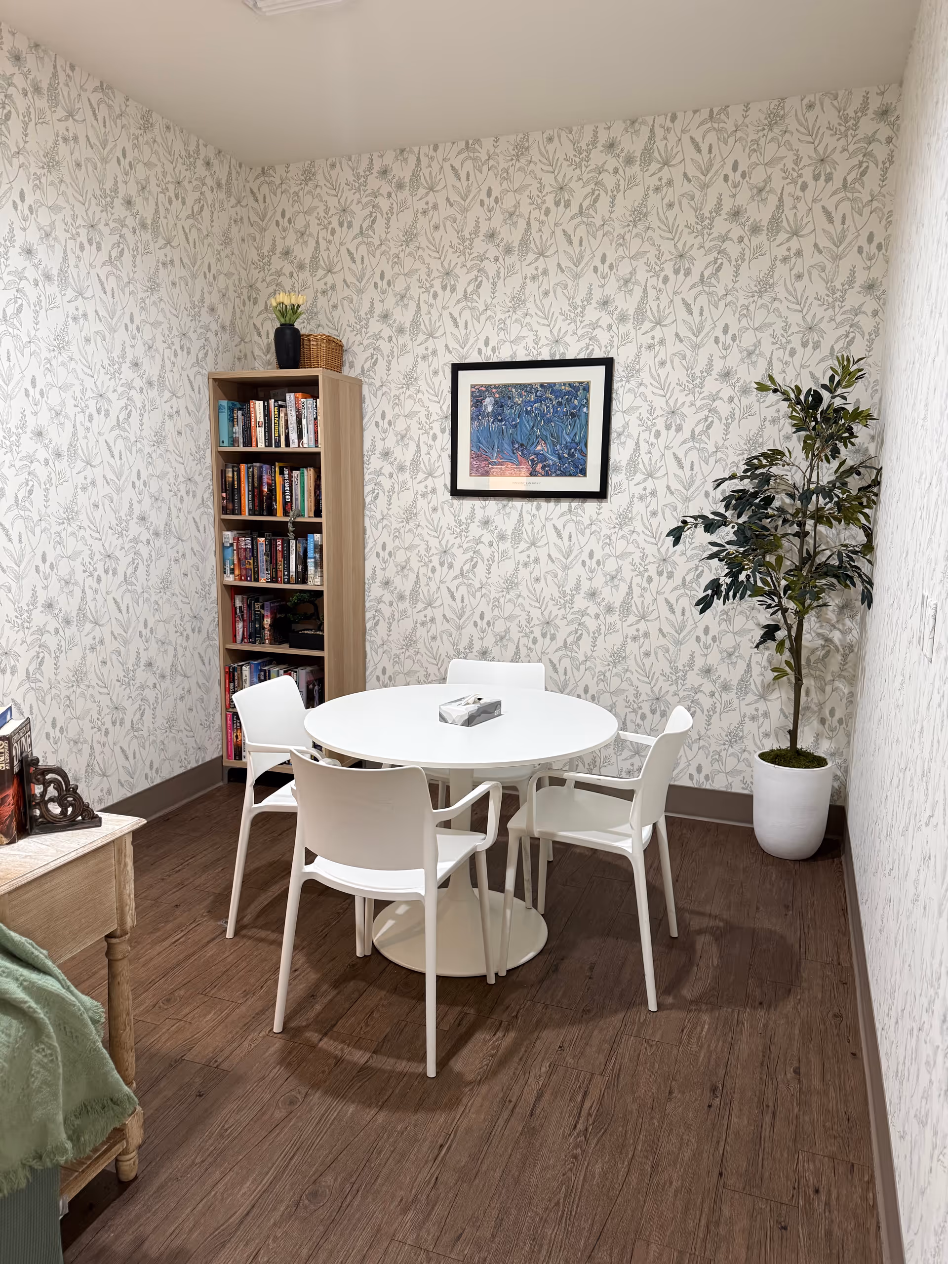 Small sitting area with a round white table surrounded by four white chairs, a bookshelf, framed art on patterned wallpaper, and a potted plant.