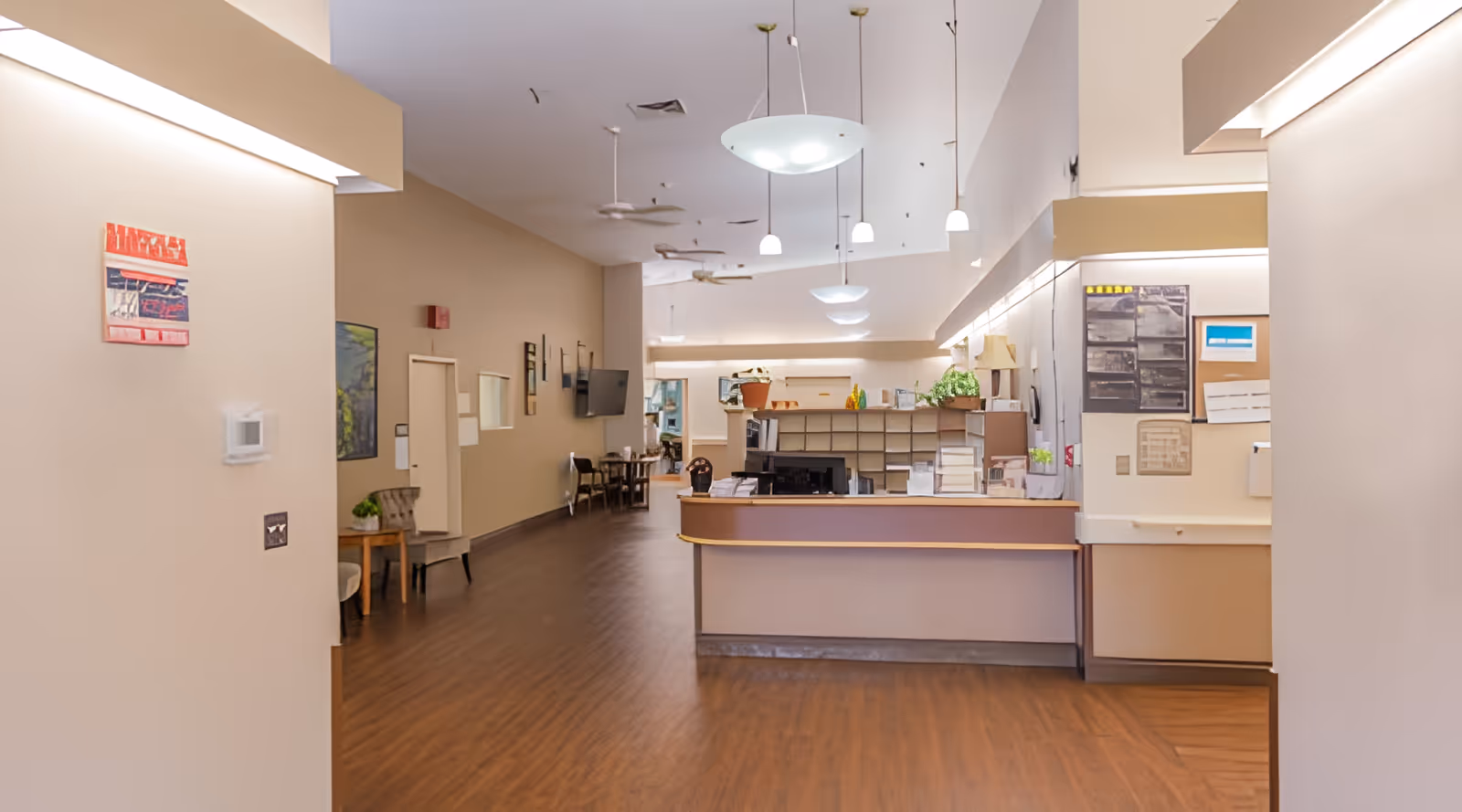 Interior view of a nursing and rehabilitation center reception area with a front desk, chairs along the wall, wall-mounted TV, and ceiling lights. The floor is wood, and the walls are painted beige with some artwork and bulletin boards.