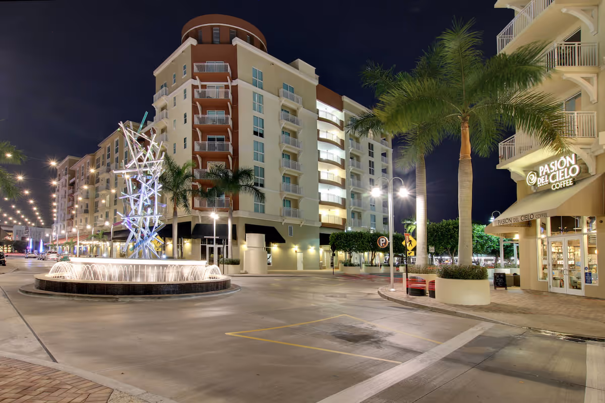 Nighttime view of a lit urban plaza with a fountain, modern multi-story building and a coffee shop under palm trees.