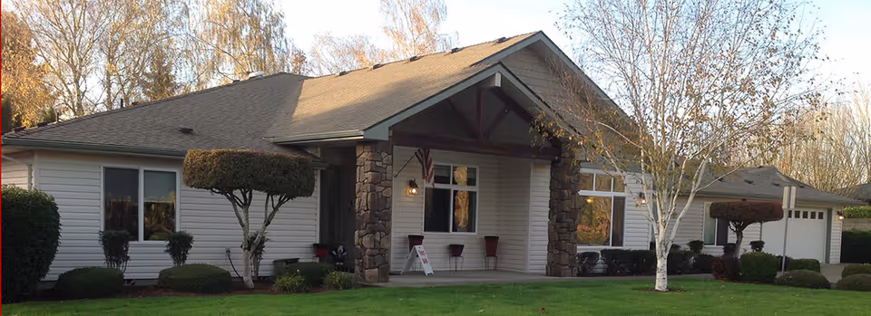 Exterior view of a single-story building with white siding and a gabled roof. The entrance features stone pillars and a small covered porch with two chairs and a sign. The surrounding area includes neatly trimmed bushes, a manicured lawn, and leafless trees under a clear sky.