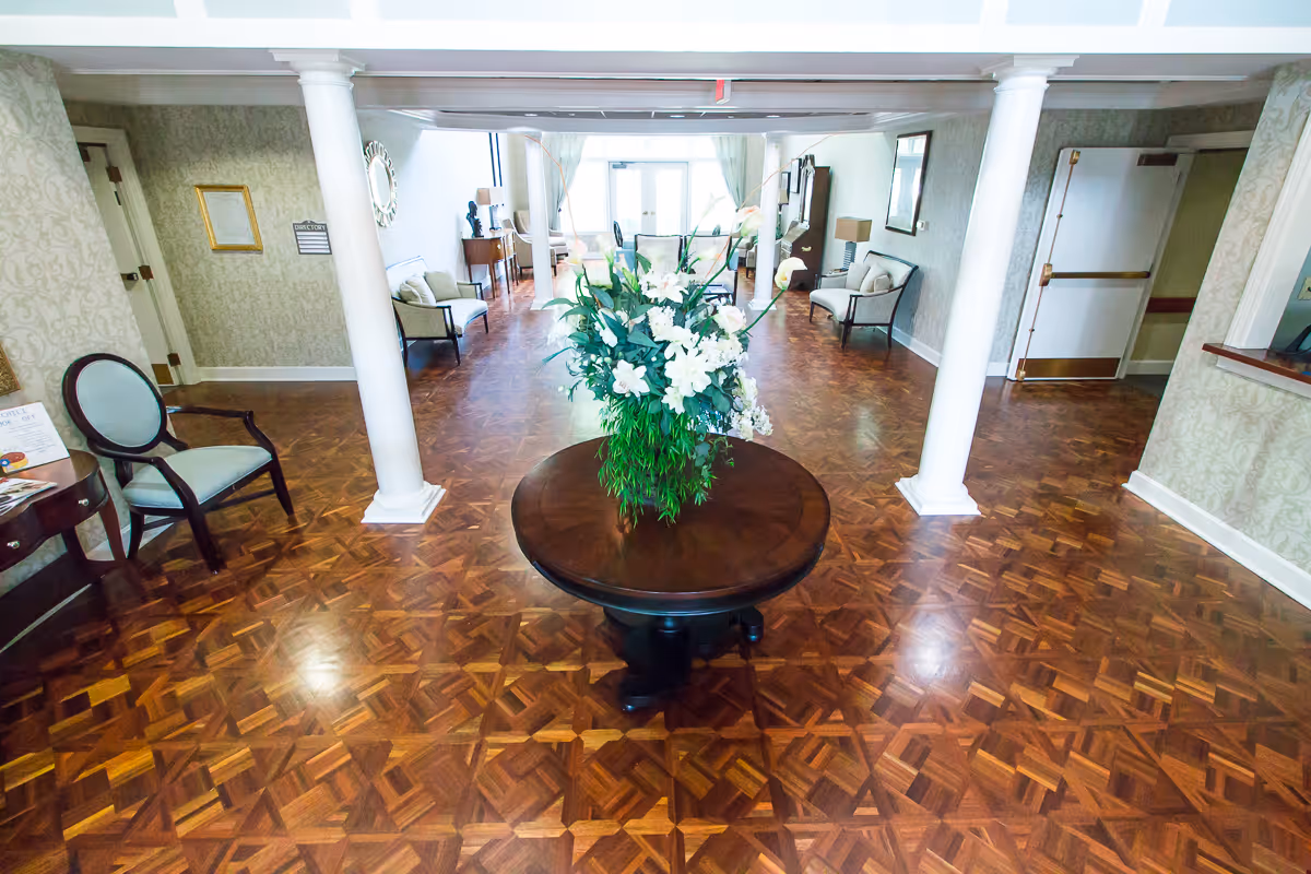 Interior view of a senior living facility lobby area with polished wooden parquet flooring, a round wooden table with a large floral arrangement in the center, white columns, cushioned chairs along the walls, and a reception desk on the right side.