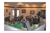 Interior view of a dining room with multiple wooden tables and chairs arranged neatly. The room is decorated with framed pictures on the walls, a chandelier hanging from the ceiling, and a planter with green foliage in the foreground.