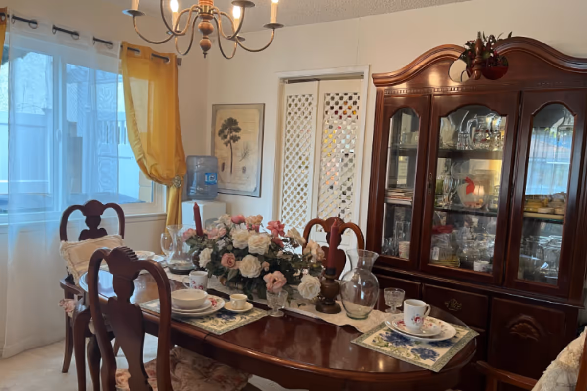 A traditional dining room with a set wooden table, floral centerpiece, china cabinet, and a window with yellow curtains.