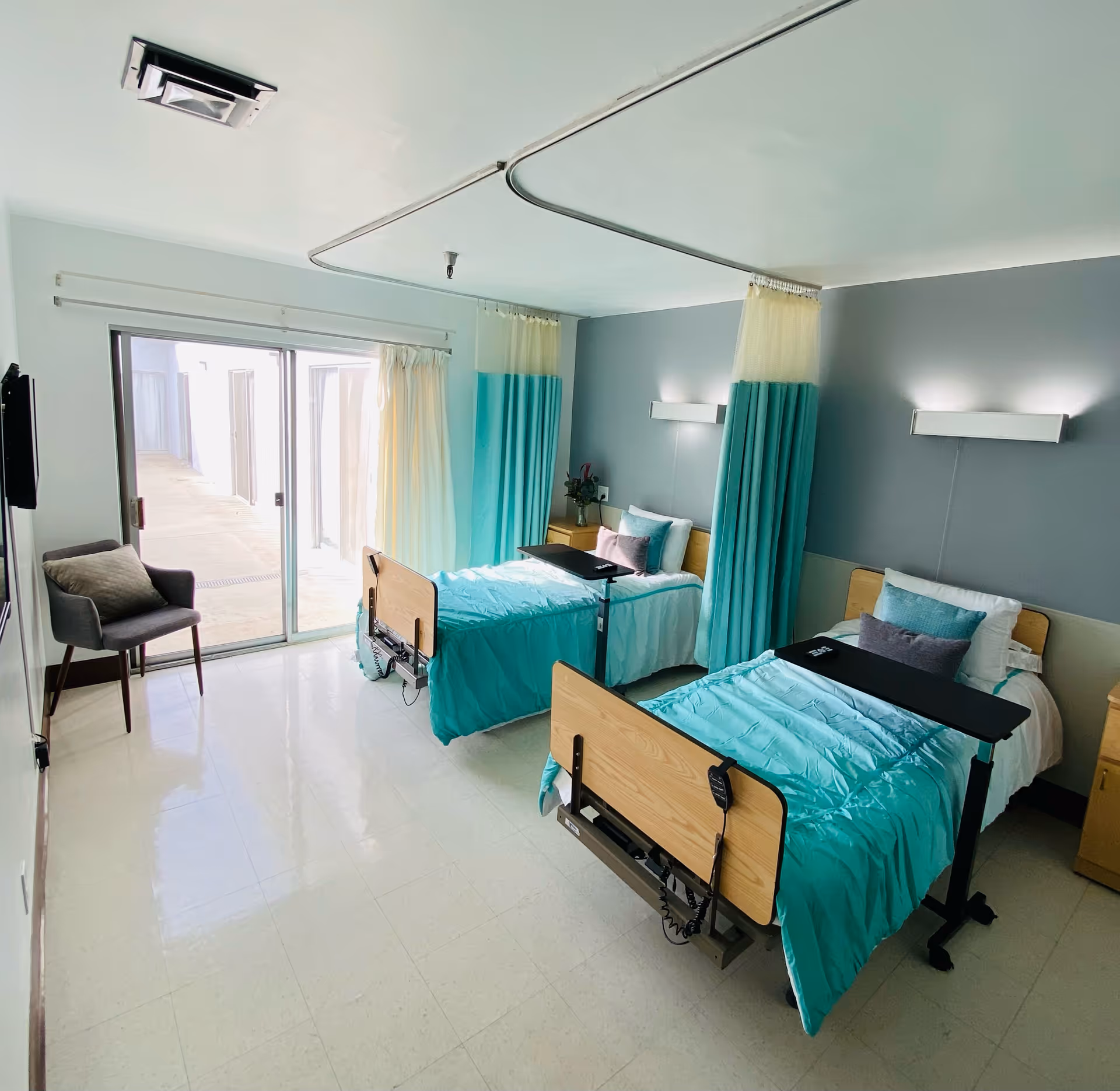 A bright and clean room with two hospital-style beds separated by turquoise and white privacy curtains. Each bed has a turquoise comforter, pillows, and an overbed table. There is a gray armchair near a sliding glass door that leads outside, and the walls are painted light gray with modern wall-mounted lights above each bed.