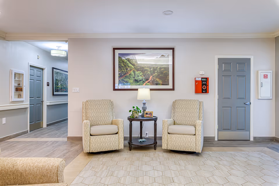 A cozy seating area in a senior living facility with two patterned armchairs placed on either side of a small round table holding a lamp, a small plant, and a framed photo. Behind the chairs is a large framed picture of a lush green forest landscape. The walls are light-colored with two gray doors and a red fire alarm box mounted on the wall. The floor has a combination of wood and tile patterns.