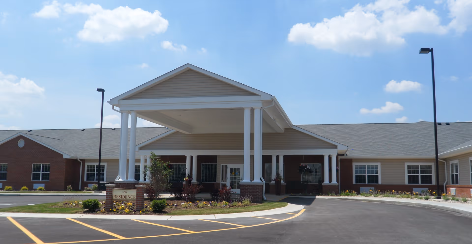 Front entrance of a single-story senior living building with a covered drop-off portico, columns, landscaping, and a paved driveway.