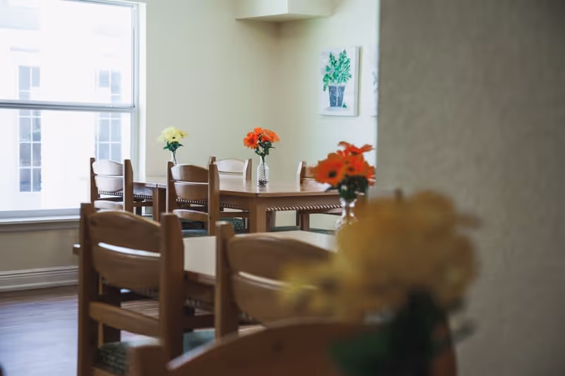 A dining area with wooden tables and chairs, each table decorated with small vases of colorful flowers. A large window lets in natural light, and a framed picture of a plant hangs on the wall.
