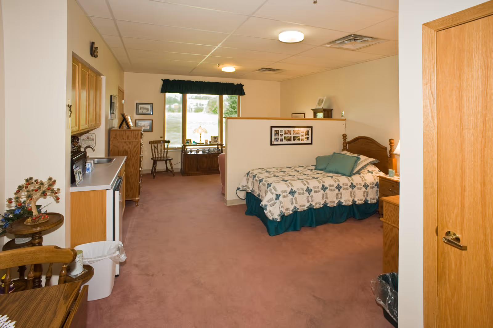 Interior of a senior living facility room featuring a bed with a patterned quilt and green pillows, a wooden nightstand with a lamp, a small kitchenette area with cabinets and a sink, a window with a dark valance, wooden furniture including chairs and a dresser, and a carpeted floor.