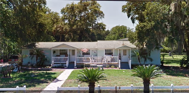 Single-story light-blue senior living facility with porch, flag, and surrounding trees.