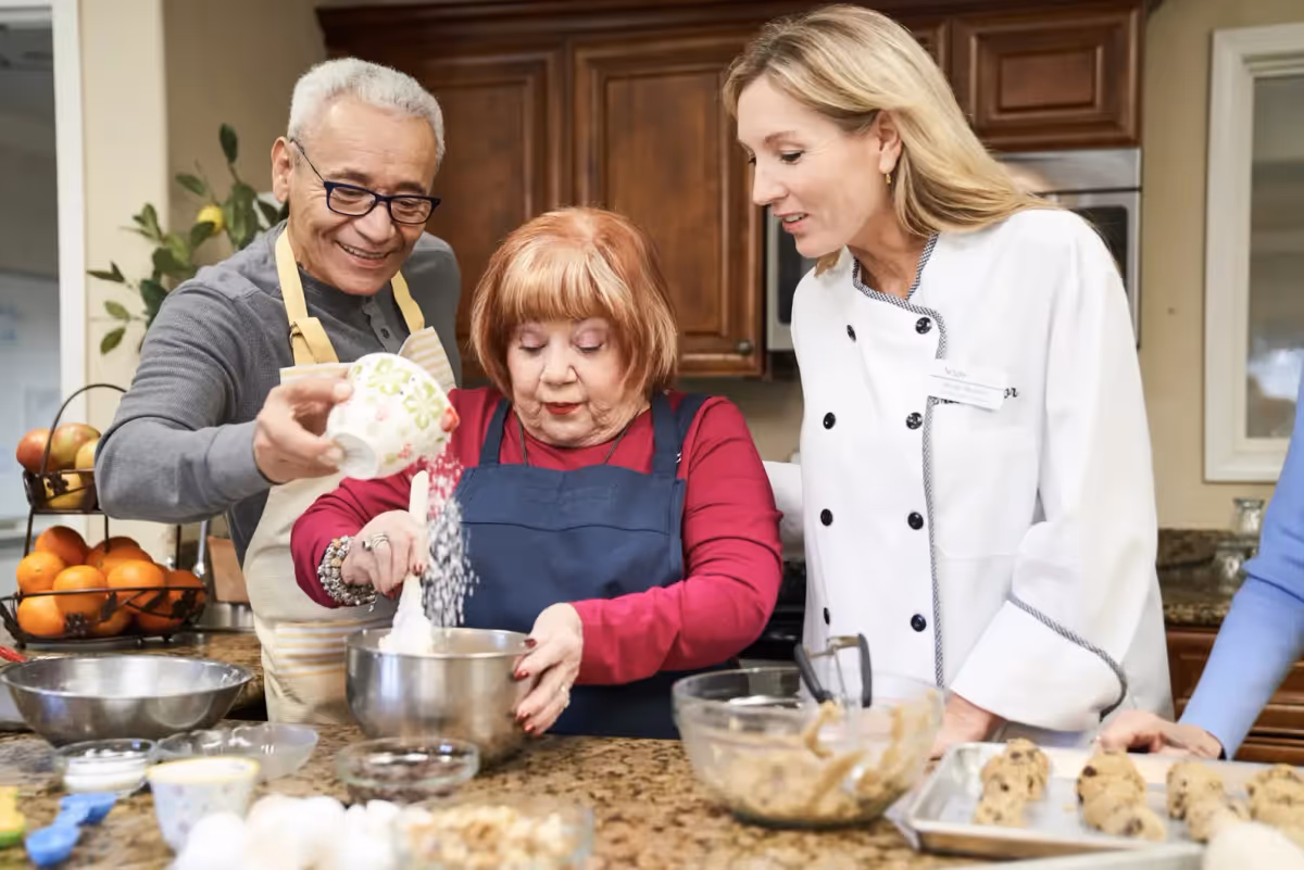 An elderly man and woman are baking together in a kitchen with the assistance of a woman in a white chef's coat. The man is pouring an ingredient from a cup into a mixing bowl held by the woman, who is stirring. Various baking ingredients and cookie dough on a tray are visible on the counter.