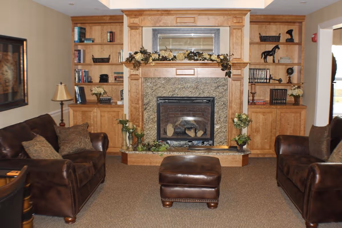 A cozy living room with two brown leather sofas facing each other, a matching ottoman in the center, and a fireplace with a wooden mantel decorated with flowers. Built-in wooden shelves flank the fireplace, holding books, decorative items, and plants. A floor lamp is positioned next to one of the sofas.