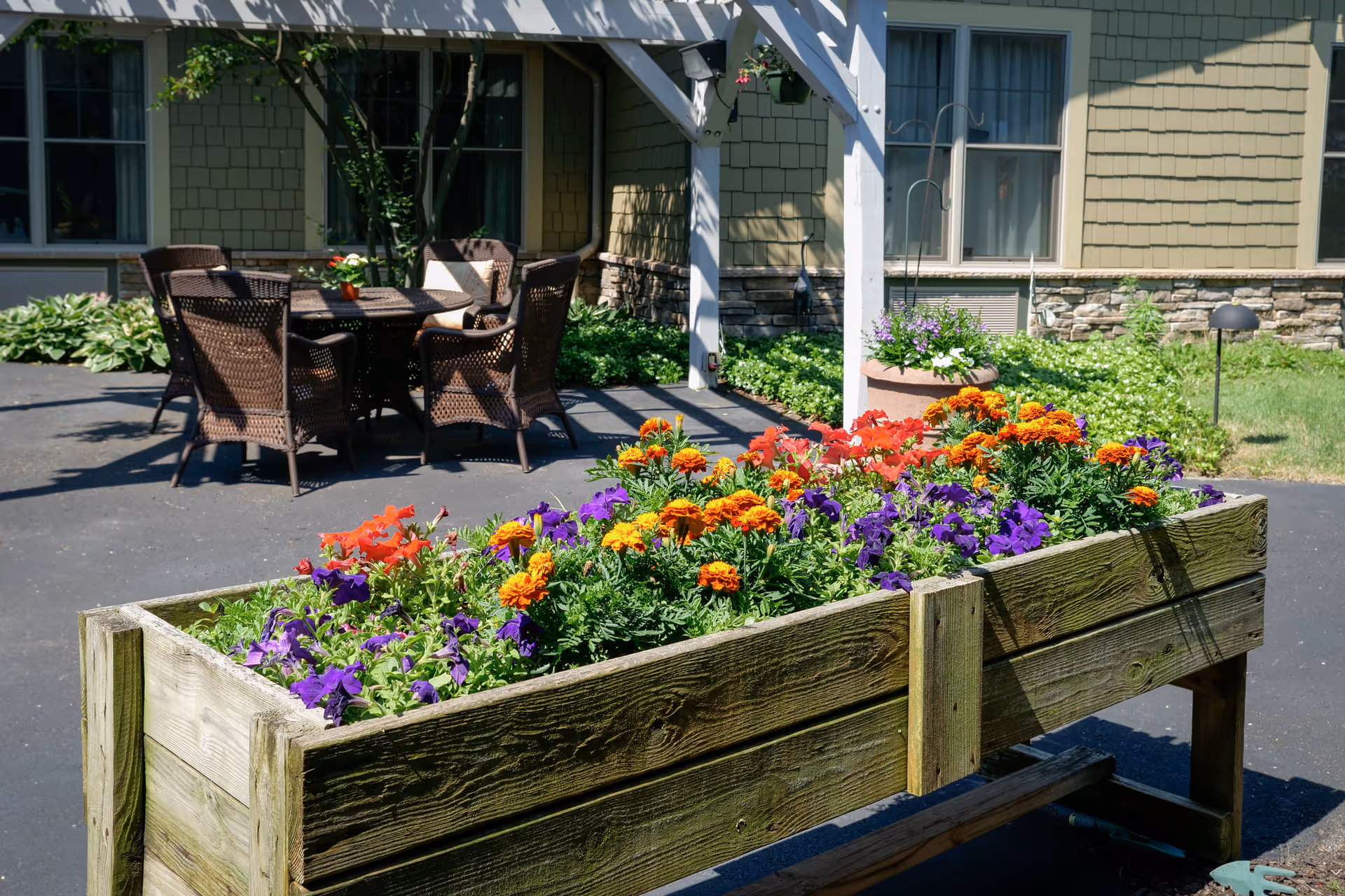 Raised wooden planter filled with colorful flowers in front of a patio seating area and the exterior of a senior living building.