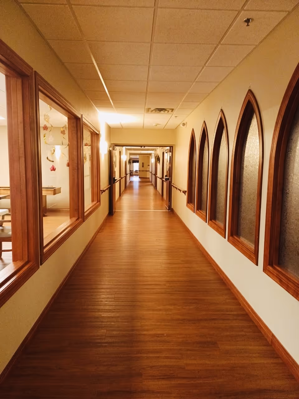 A long, well-lit hallway with wooden flooring and beige walls. On the left side, there are large windows looking into a room with tables and chairs. On the right side, there are arched windows with frosted glass. The ceiling has recessed lighting and a drop ceiling design.