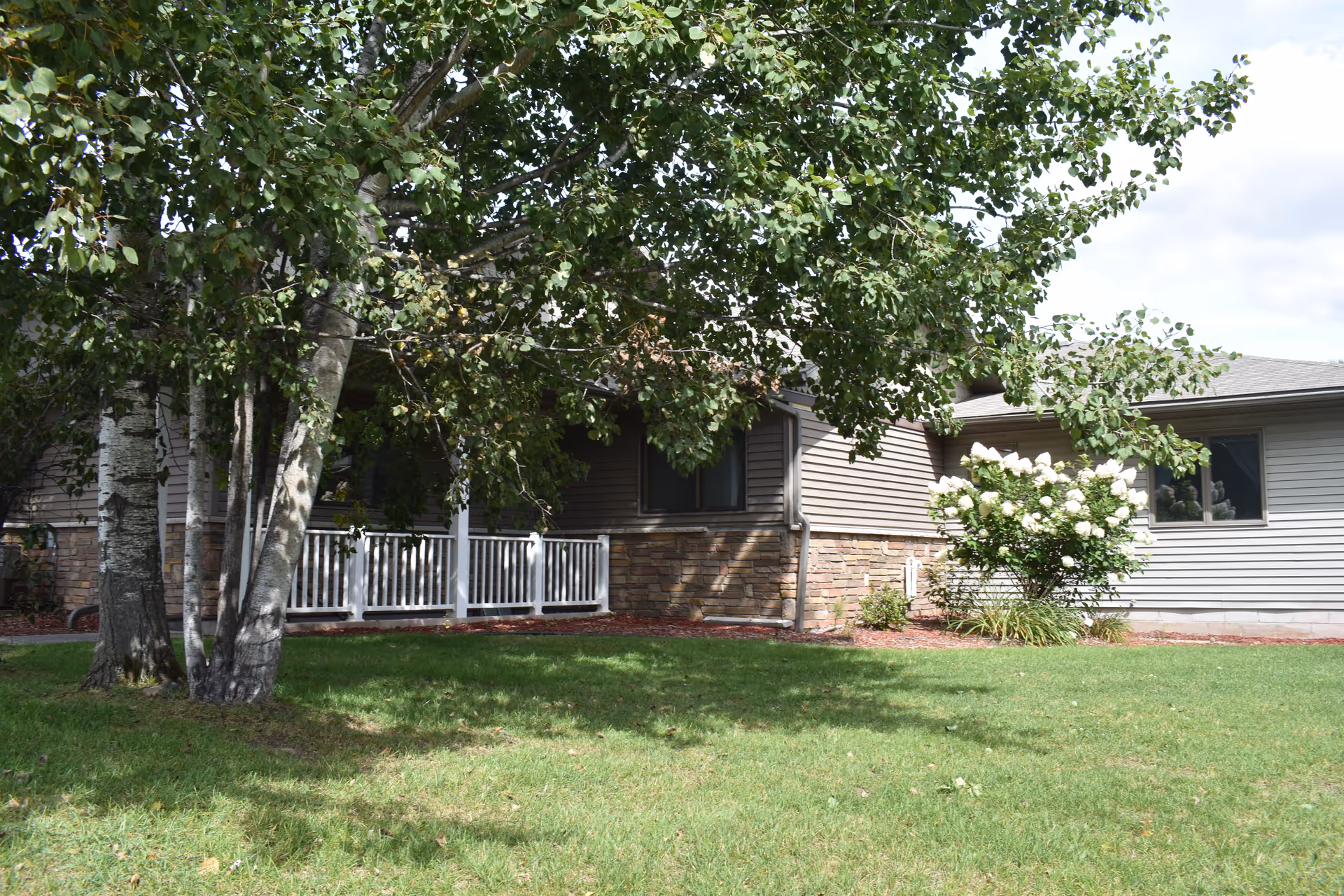 Exterior view of a single-story building with beige siding and stone accents, partially shaded by a large leafy tree in the foreground. There is a white railing along a small porch area and a flowering bush with white blooms near the building. The lawn is green and well-maintained under a partly cloudy sky.